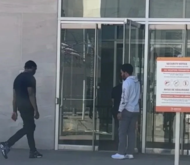 Three people near a glass entrance door of a building, with one person entering and two others standing nearby.