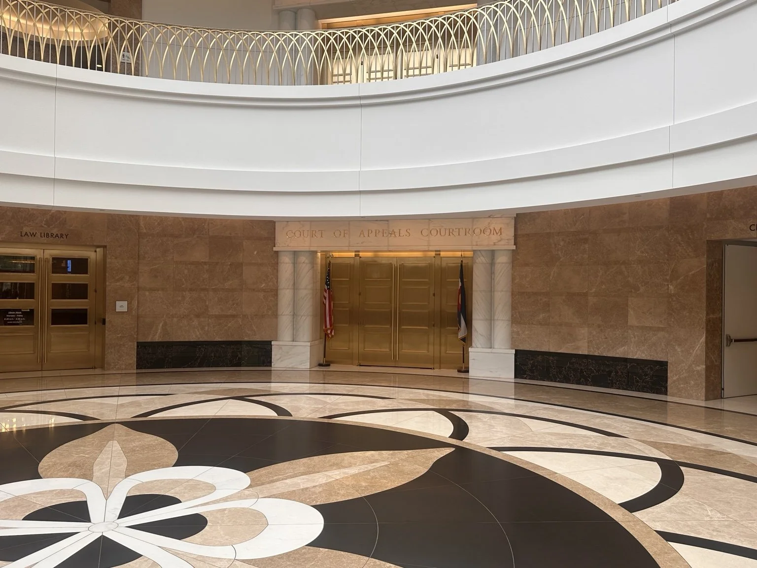 Interior view of a courthouse with marble flooring, gold doors labeled 'Court of Appeals Courtroom,' and flags beside the doors. The upper level has a curved, decorative white railing.