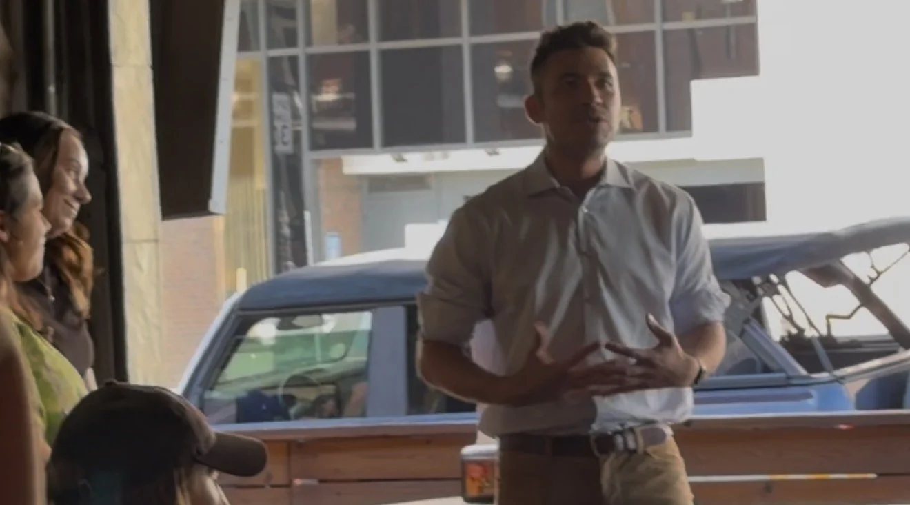 A man in a white shirt standing and speaking in front of a group inside a building with large windows.