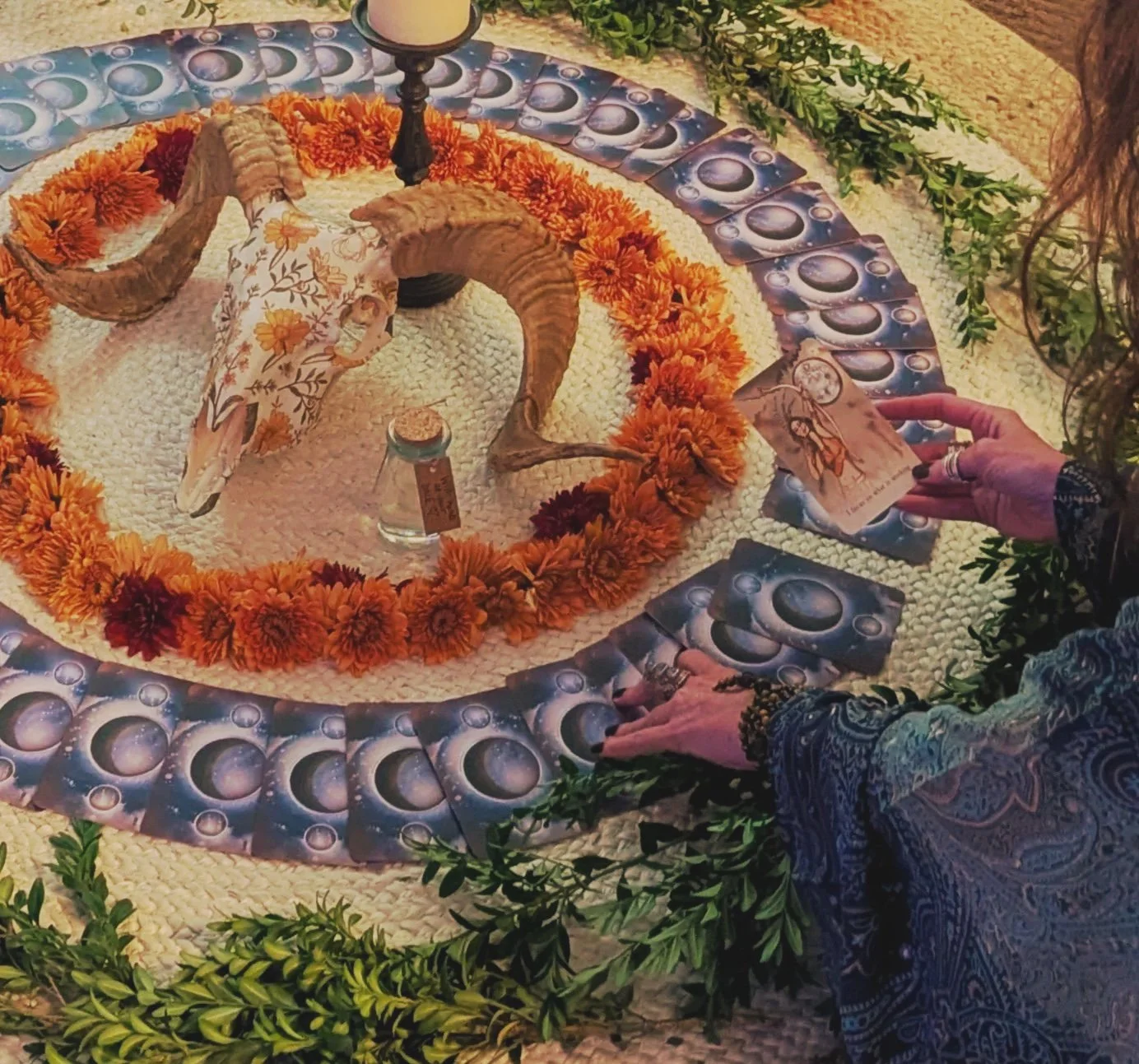 A altar with a ram skull at the center, surrounded by a wreath of orange and red flowers, with tarot cards laid on the surface, and a person holding one tarot card.