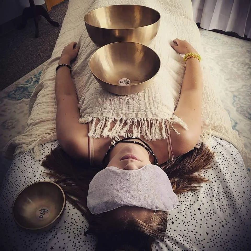A woman lying on her back on a massage table with Himalayan bowls on her chest and stomach, and a blanket over her. She wears an eye covering.