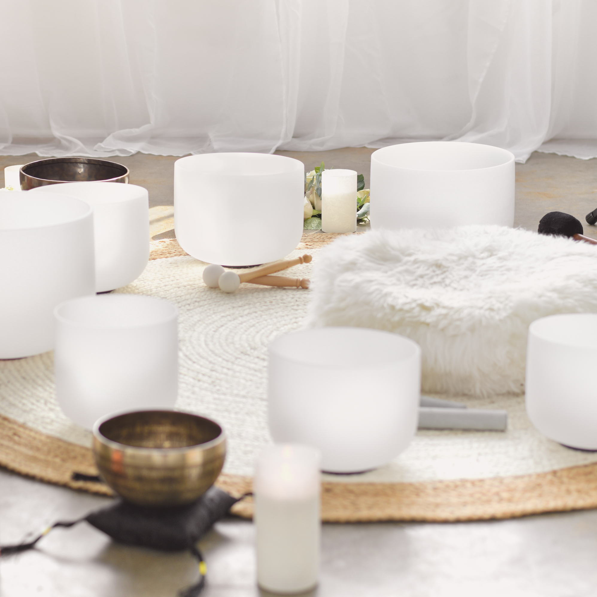 A collection of crystal and metal singing bowls and candles arranged on a woven rug, with a white fluffy cushion in front.