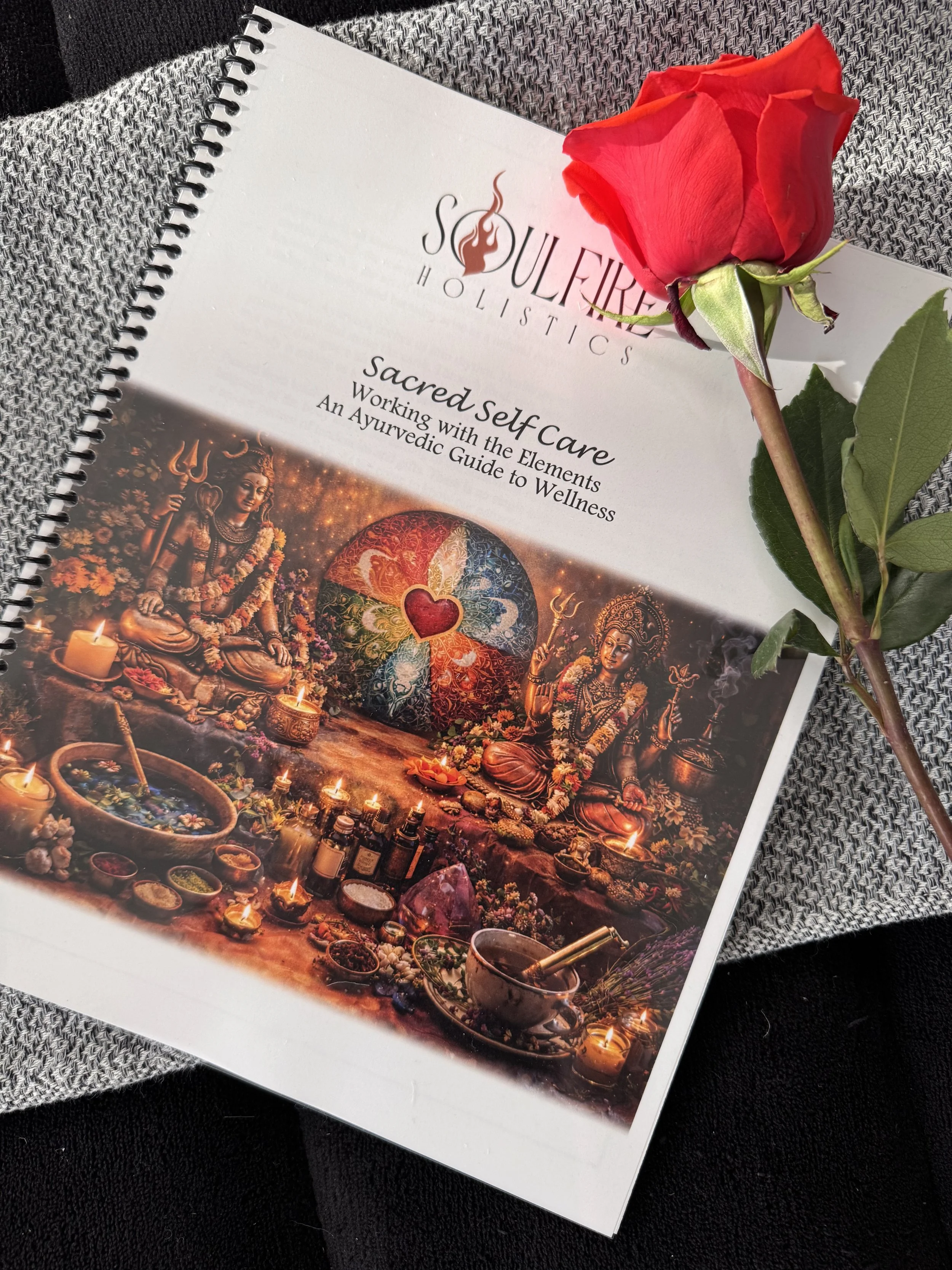 A spiral notebook titled "Sacred Self Care" with the subtitle "Working with the Elements - An Ayurvedic Guide to Wellness". The cover features an image of an Indian altar with statues, candles, herbs, and a rainbow-colored chakra symbol with a heart at the center. A pink rose is placed on the notebook.