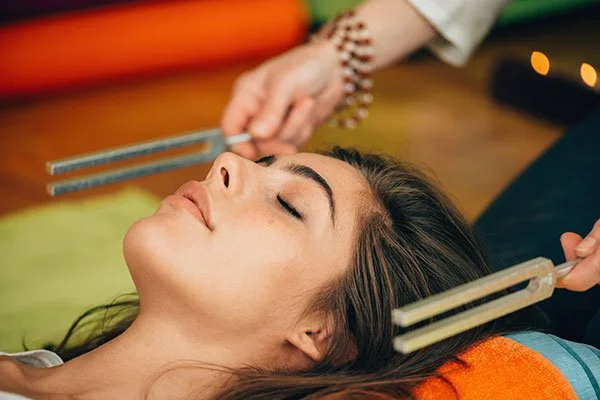 A woman lying down with her eyes closed during a sound healing treatment, with a practitioner using tuning forks on both sides of her head.