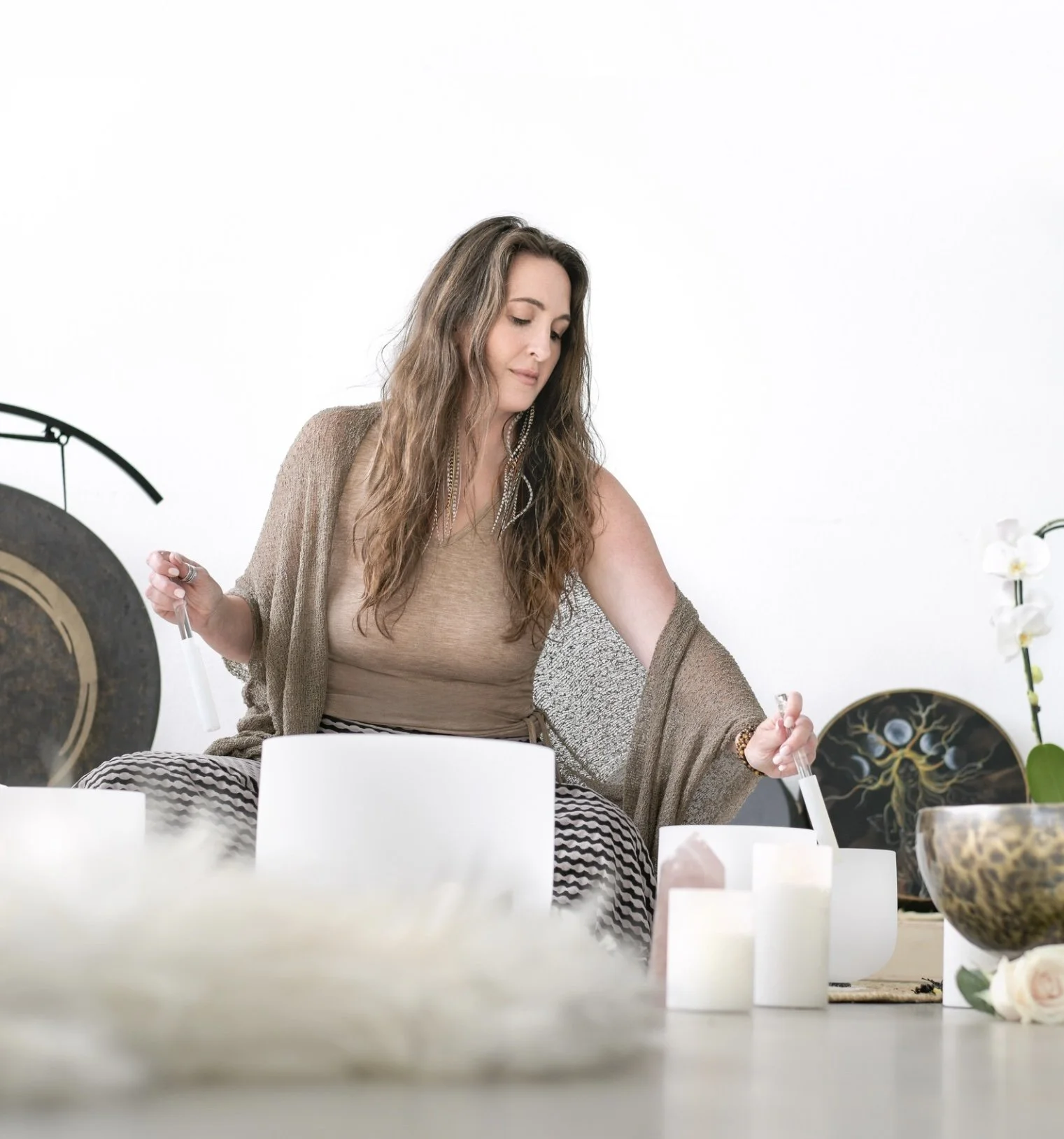 A woman with long wavy hair sitting cross-legged on the floor, near two white crystal singing bowls mallets, surrounded by candles, a large drum, and crystals, with a white background.