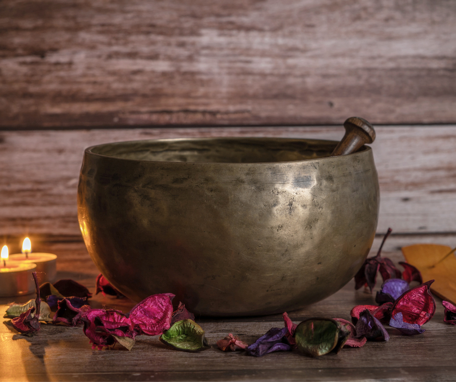 A brass singing bowl with a wooden striker resting inside, surrounded by colorful dried flower petals and a lit candle on a wooden surface against a wooden wall background.