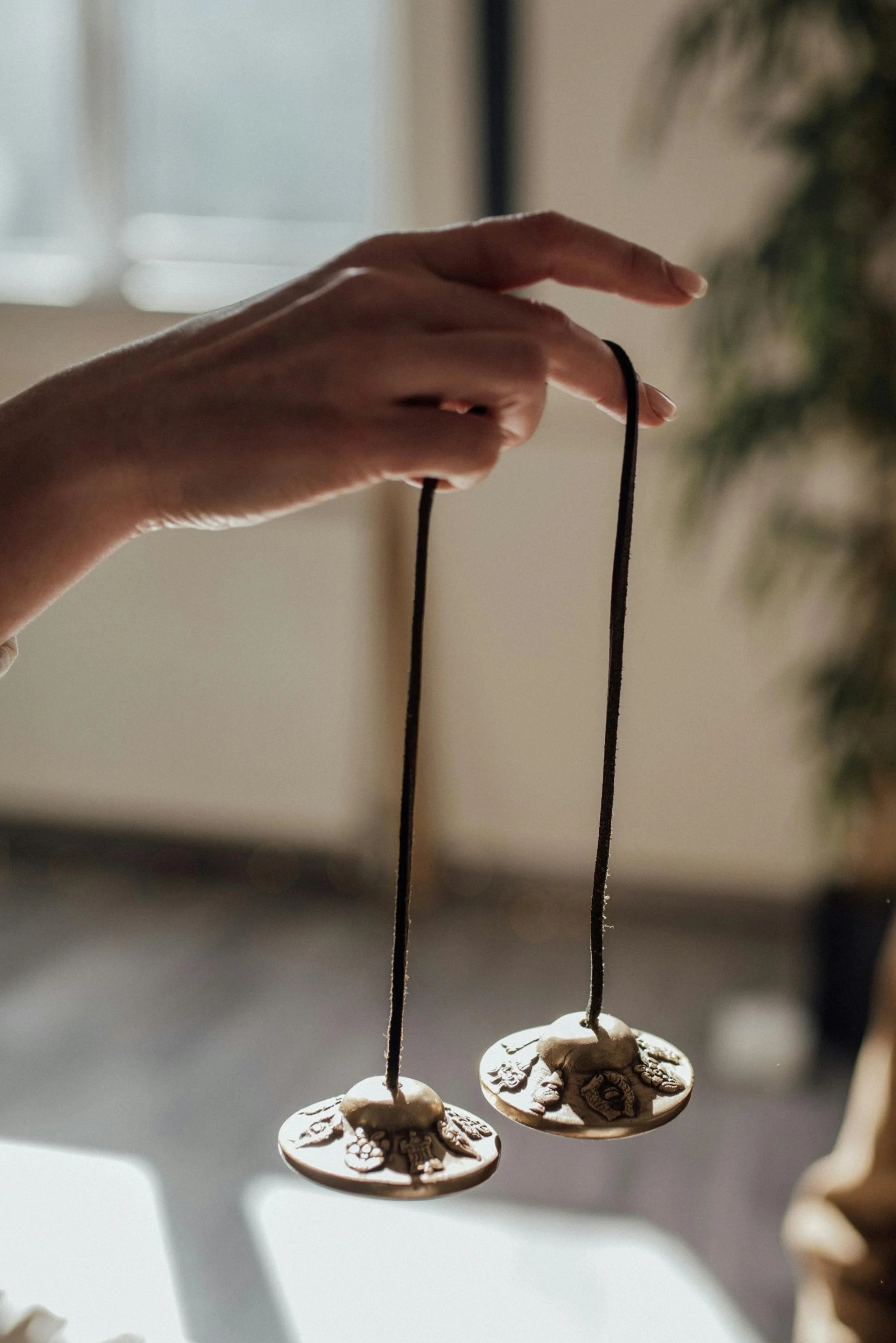 Close-up of a hand holding tingshas hanging from black strings, with a blurred indoor background.