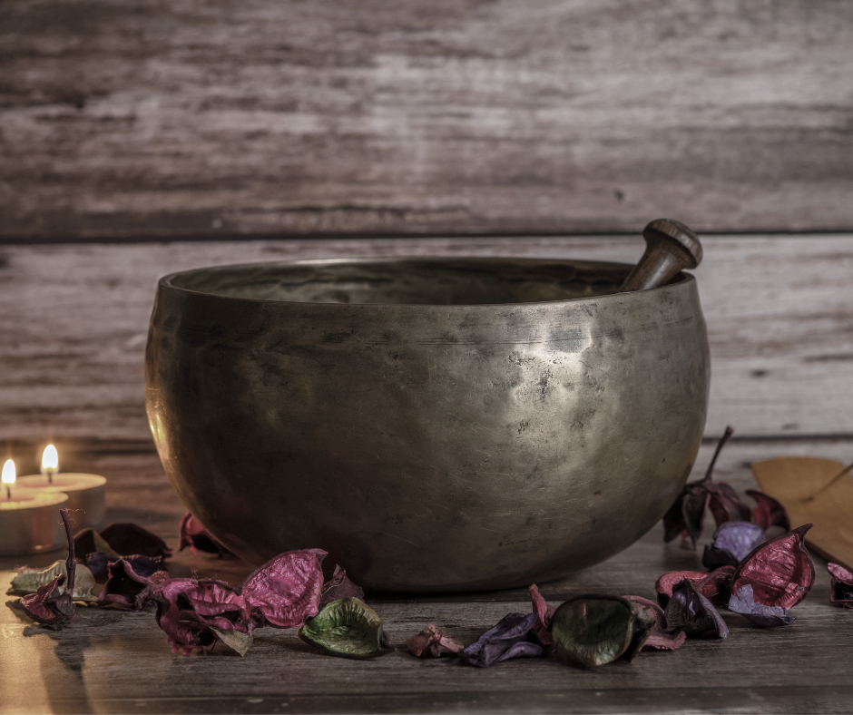 A singing bowl with a mallet inside, surrounded by dried flower petals and lit tea light candles on a wooden surface with a wooden wall background.