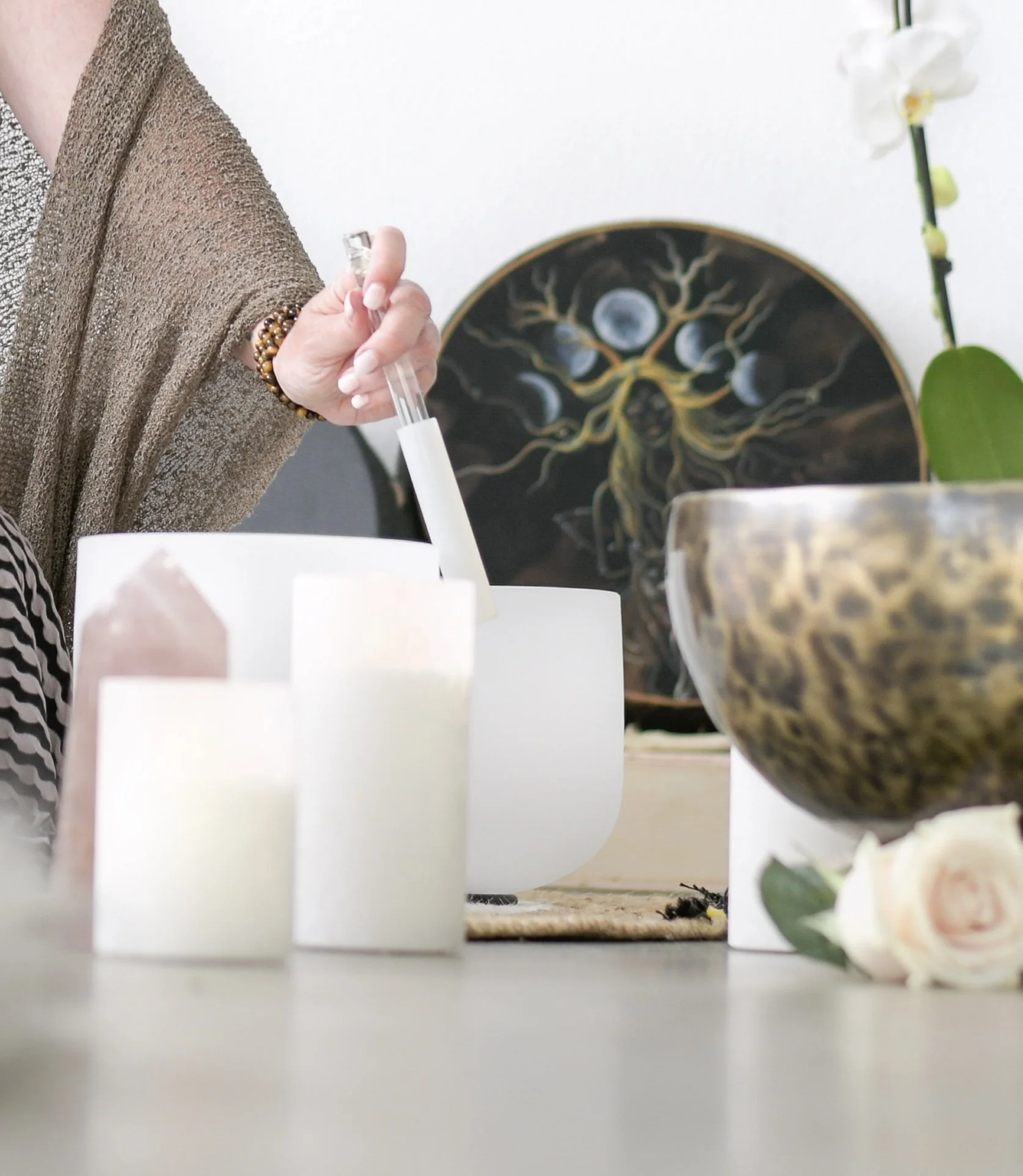 A person holding a mallet over a white crystal singing bowl, with candles and a flower arrangement on the floor. An artistic painting of a woman with moons is painted on a nearby shamanic drum.