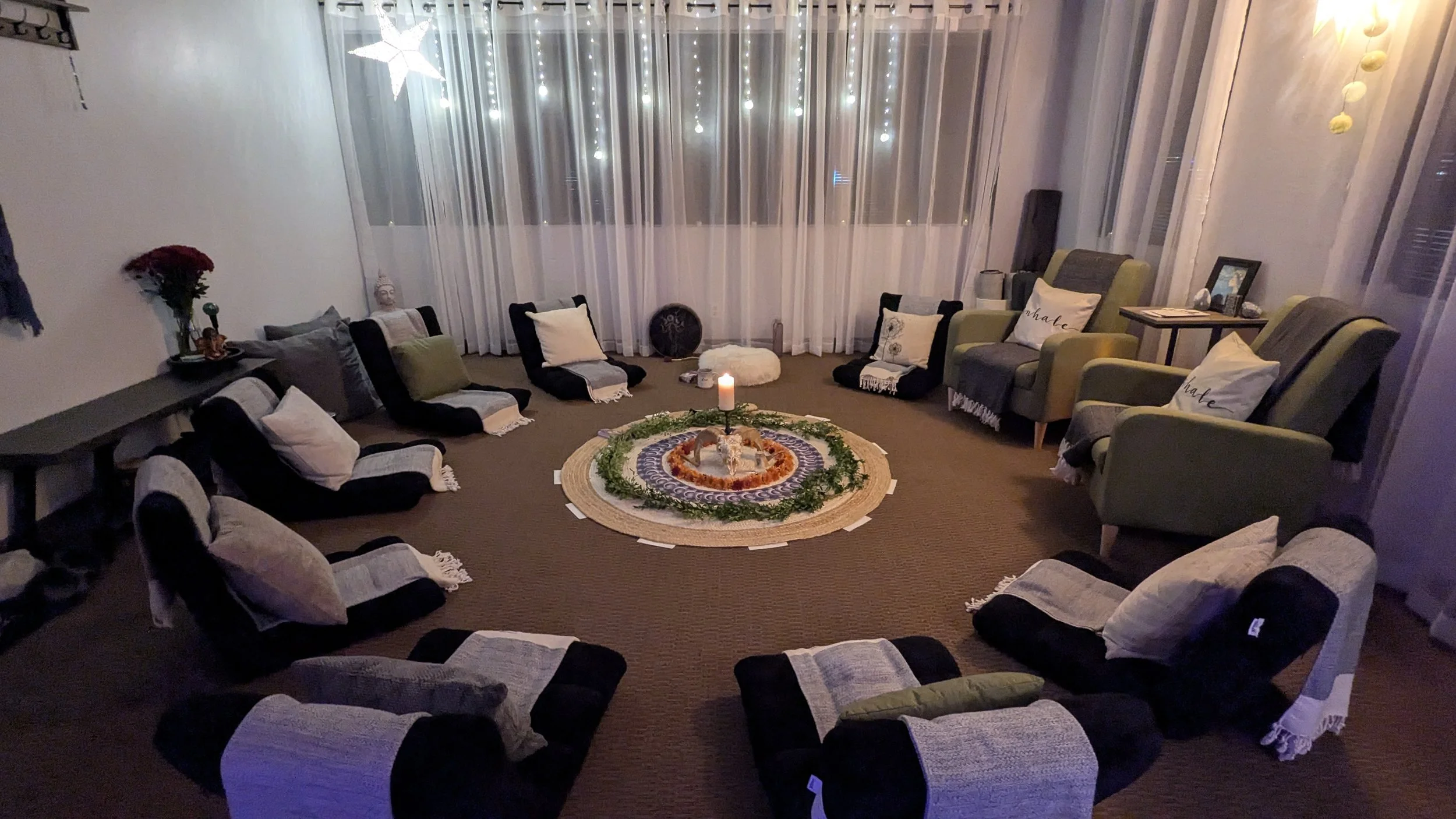 A cozy living room with a circular arrangement of floor chairs with cushions, a central candle, and decorative items, illuminated by string lights and a hanging star-shaped light.