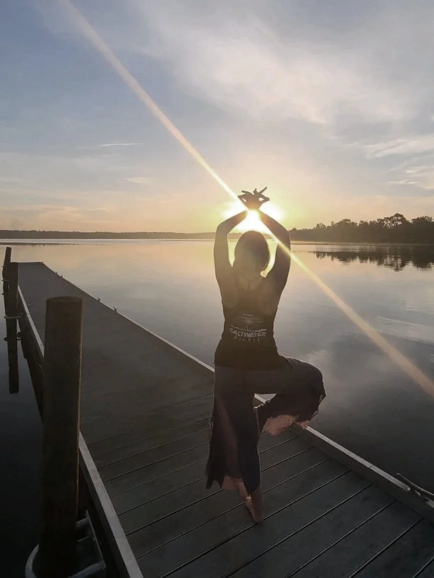 Person performing a yoga pose in a tree shape on a dock at sunset over a calm body of water.