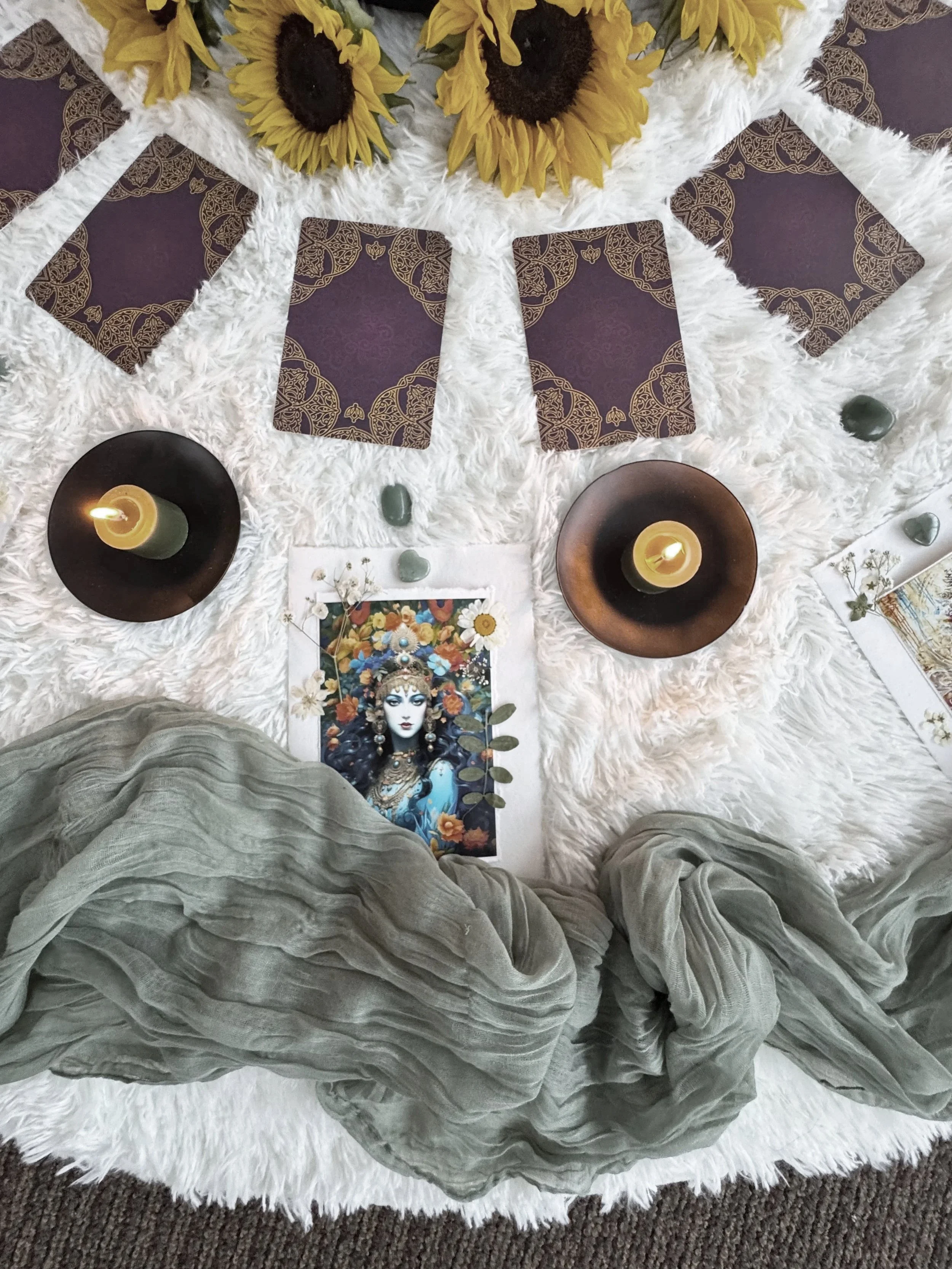 A spiritual or ritual setup on a white fluffy surface with oracle cards, candles, a picture of a goddess with flowers, sunflower decorations, and green stones.