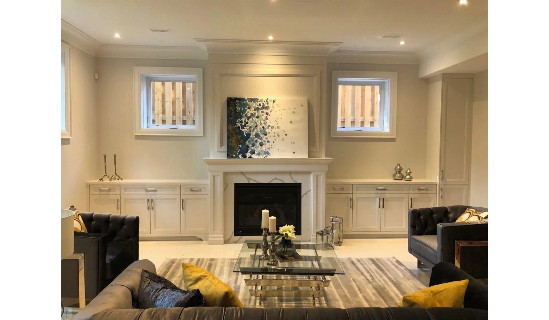 Living room with beige walls, white built-in cabinets, a fireplace with art above, and two small windows. There are two black armchairs, a sofa with yellow and black pillows, a glass coffee table with candles and flowers, and decorative objects on the cabinets.