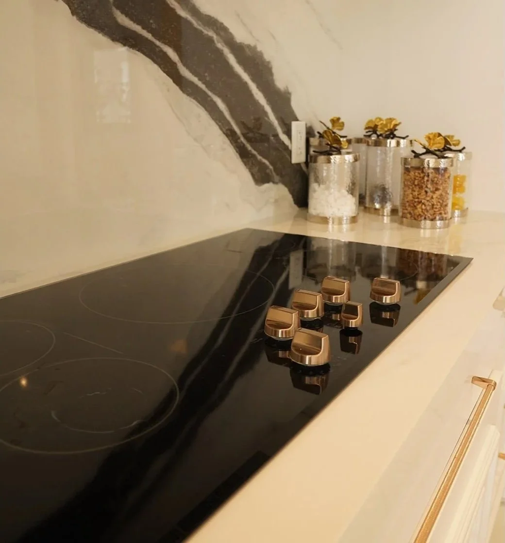 Close-up of a black electric stovetop with seven brown control knobs, situated on a cream-colored countertop in a kitchen. In the background, there are glass jars with yellow and brown decorative lids, containing various dried ingredients, arranged in front of a marble backsplash with black, white, and gold accents.