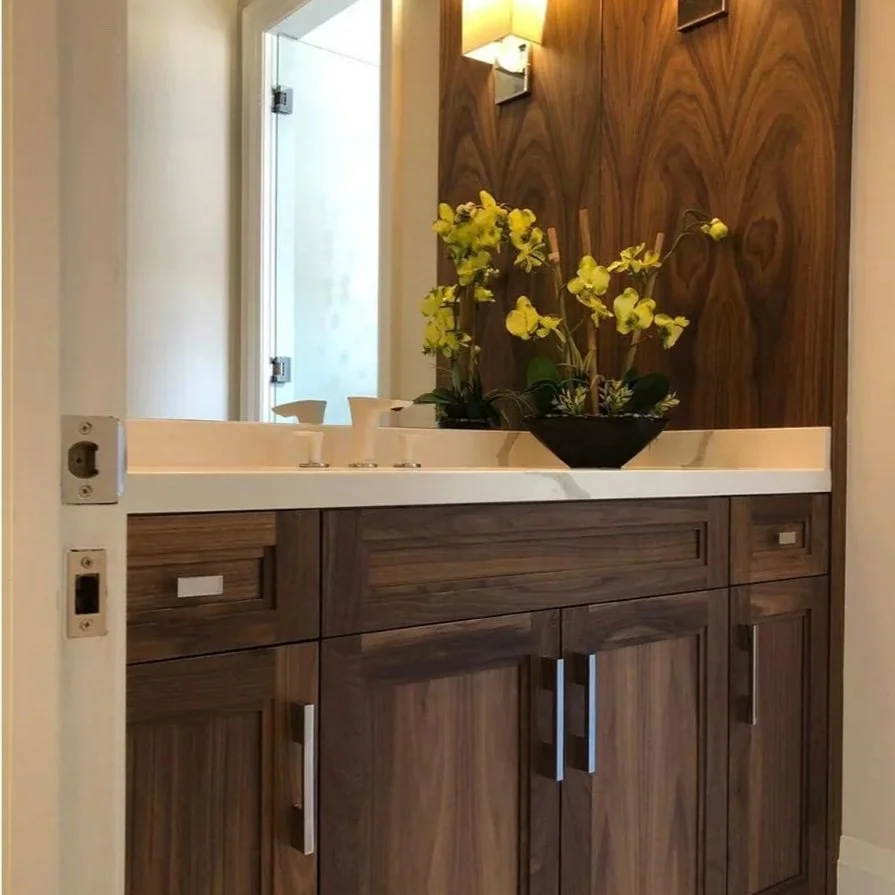 Wooden bathroom vanity with a white marble countertop, a black flower vase with yellow orchids, a mirror, and a wall-mounted light fixture.