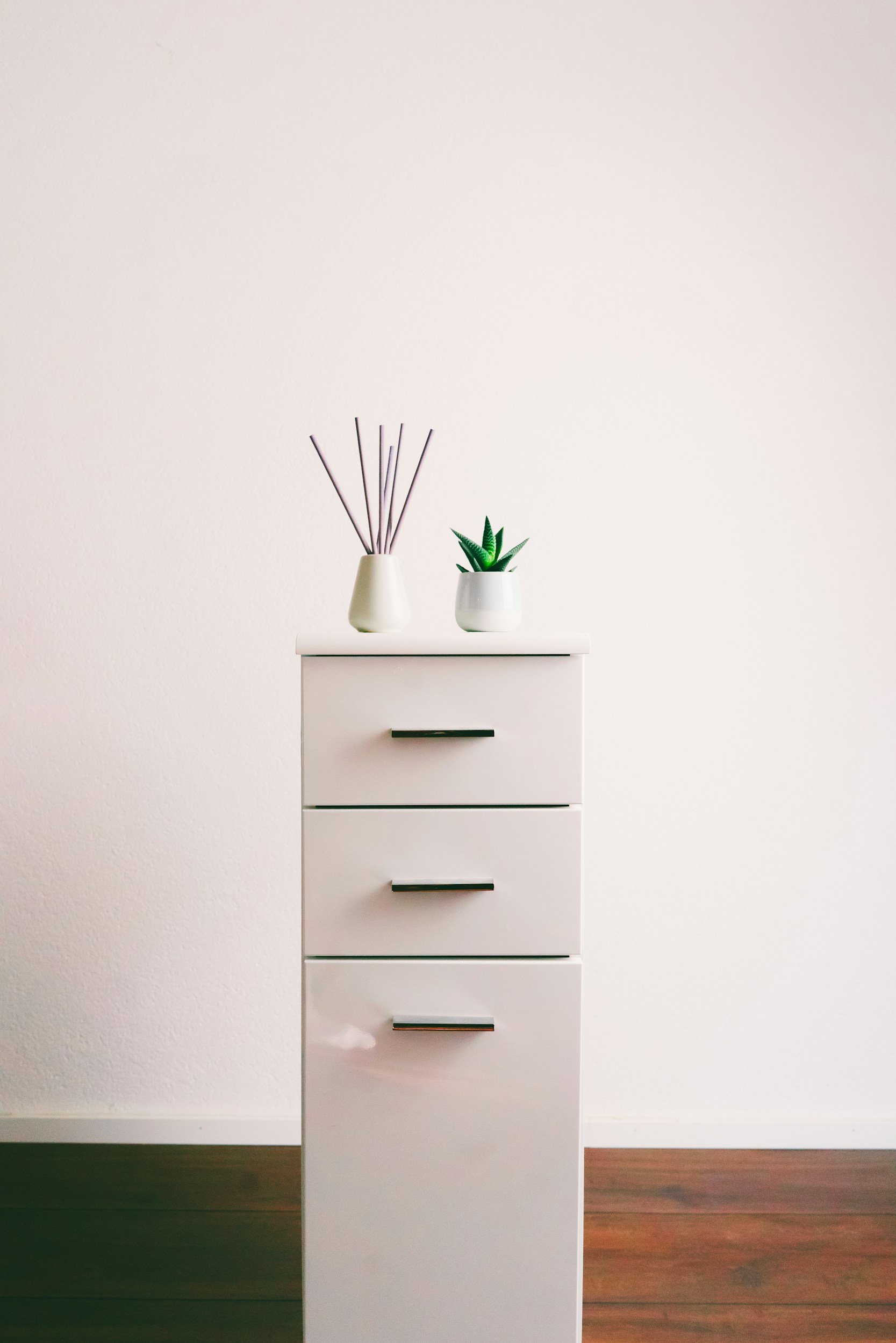 A white three-drawer dresser with black handles, topped with a white diffuser and a small potted green plant, against a plain white wall and brown wooden floor.