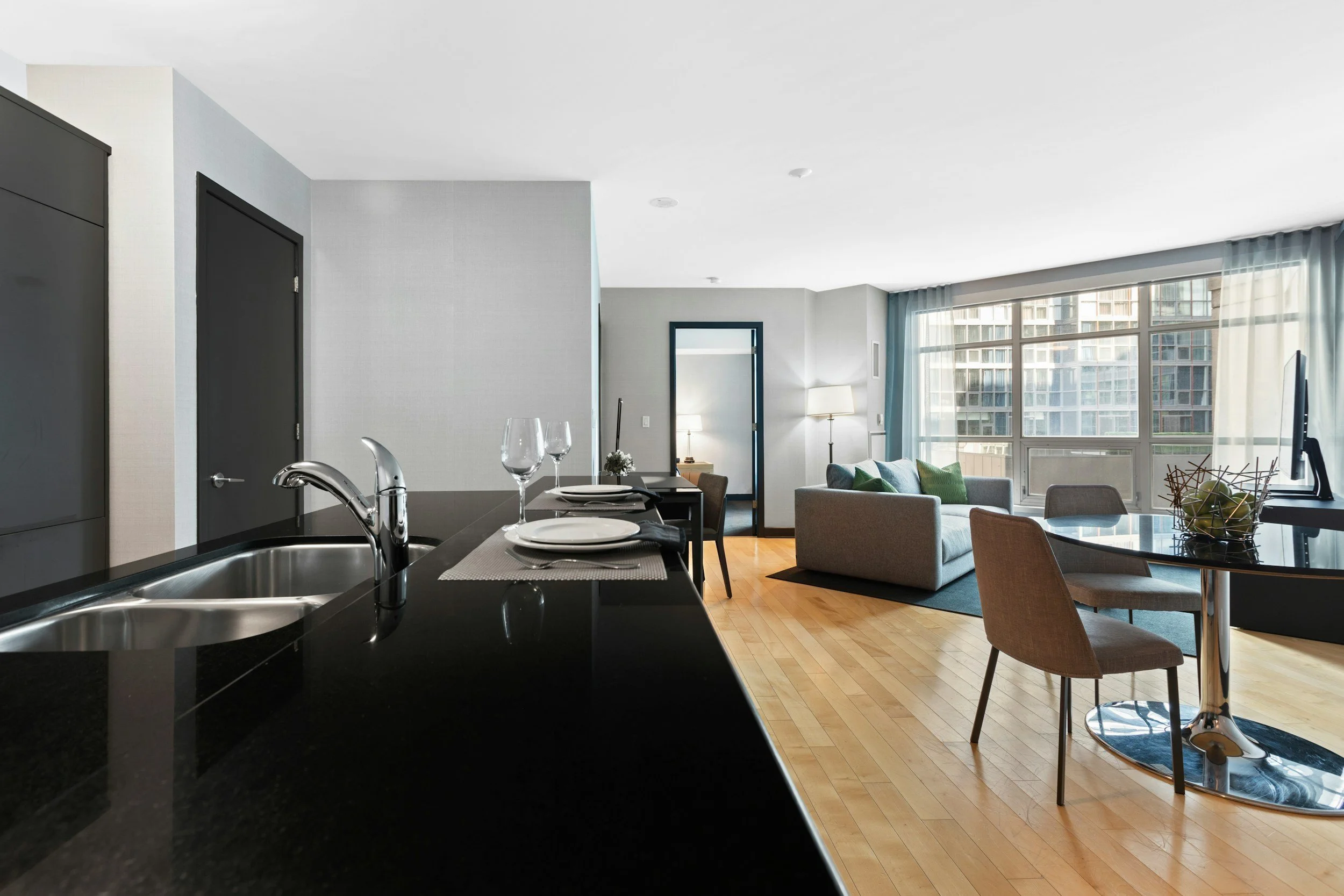 Modern open-concept apartment living area with a black kitchen counter, dining table set with plates and glasses, and a seating area with a sofa, chairs, and large windows letting in natural light.