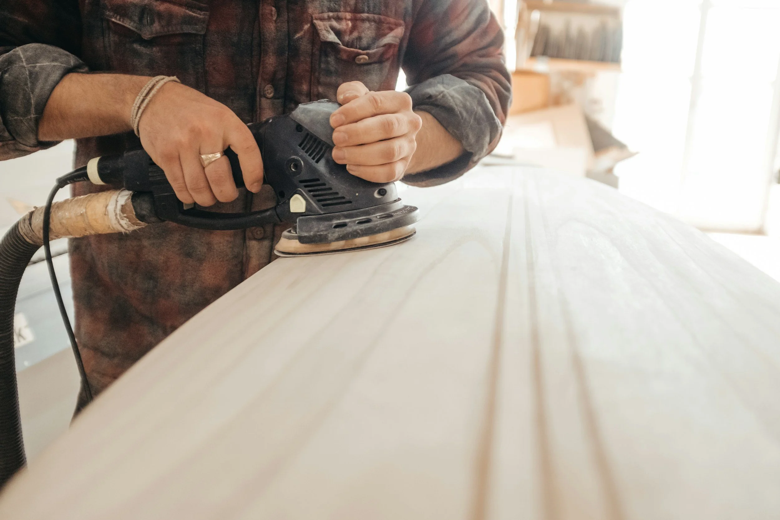 Person sanding a long wooden board with a handheld electric sander in a woodworking workshop.