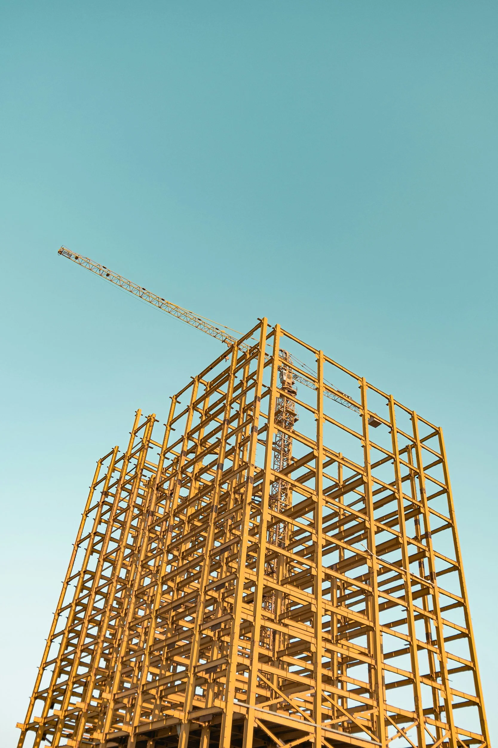 Construction site with a yellow steel framework and a tall crane against a clear blue sky.