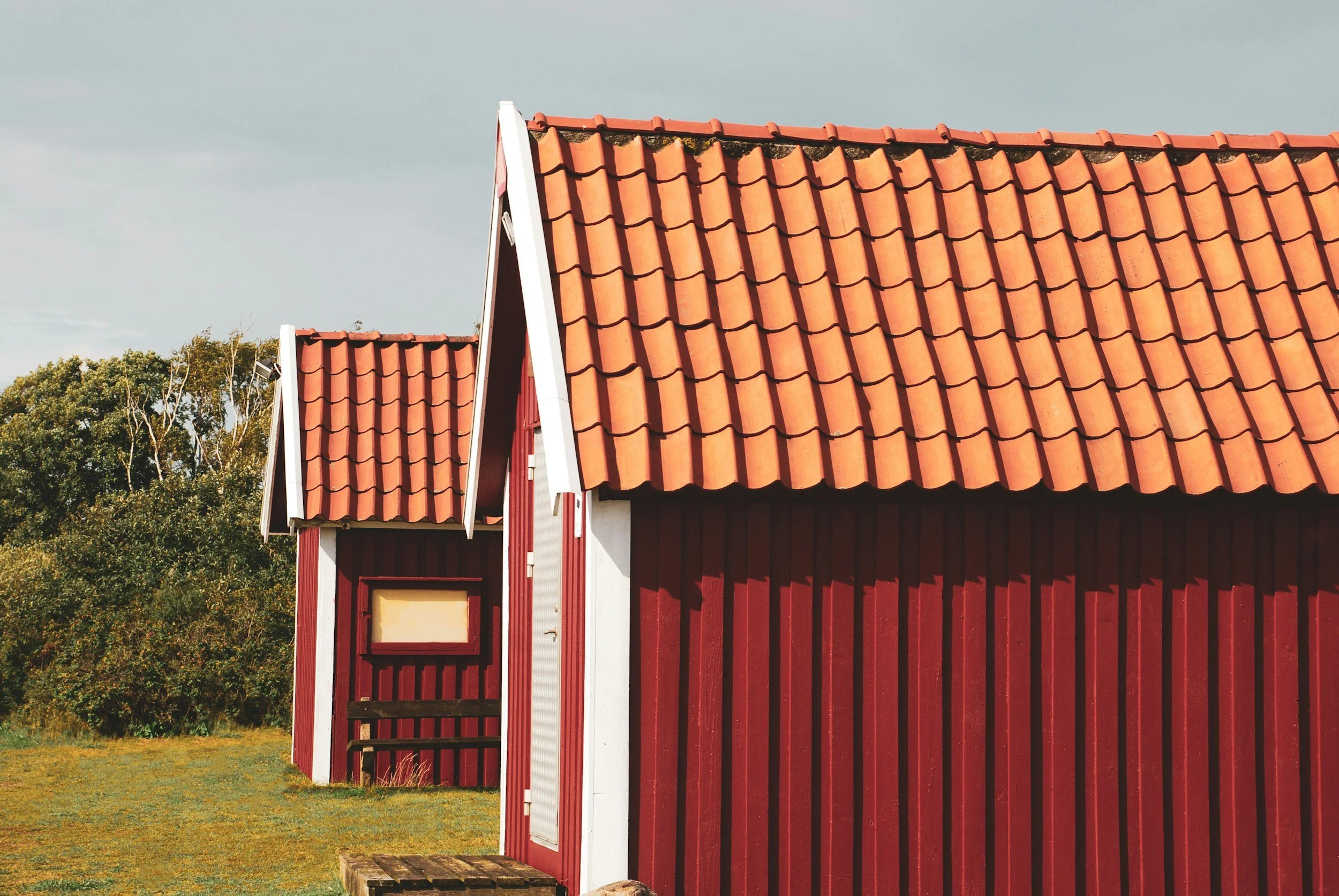 Red wood cabins with orange tiled roofs in a grassy area with trees in the background under a cloudy sky.