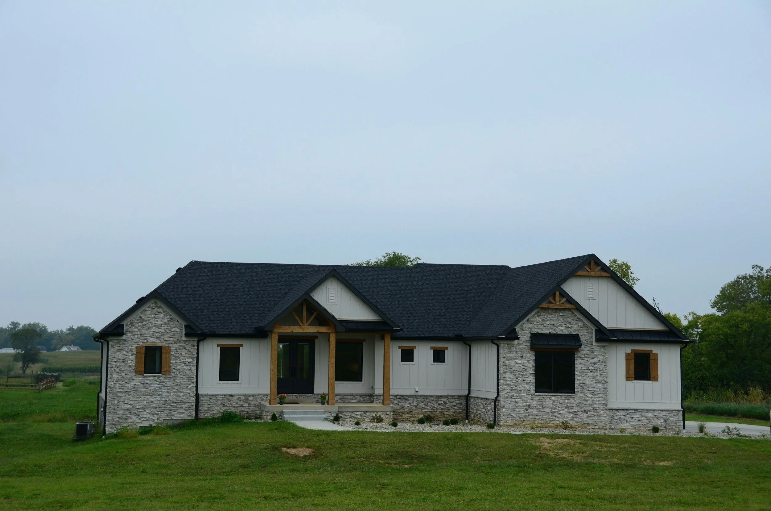 A modern one-story house with a dark shingle roof, white vertical siding, and gray stone accents, situated on a grassy lot under an overcast sky.