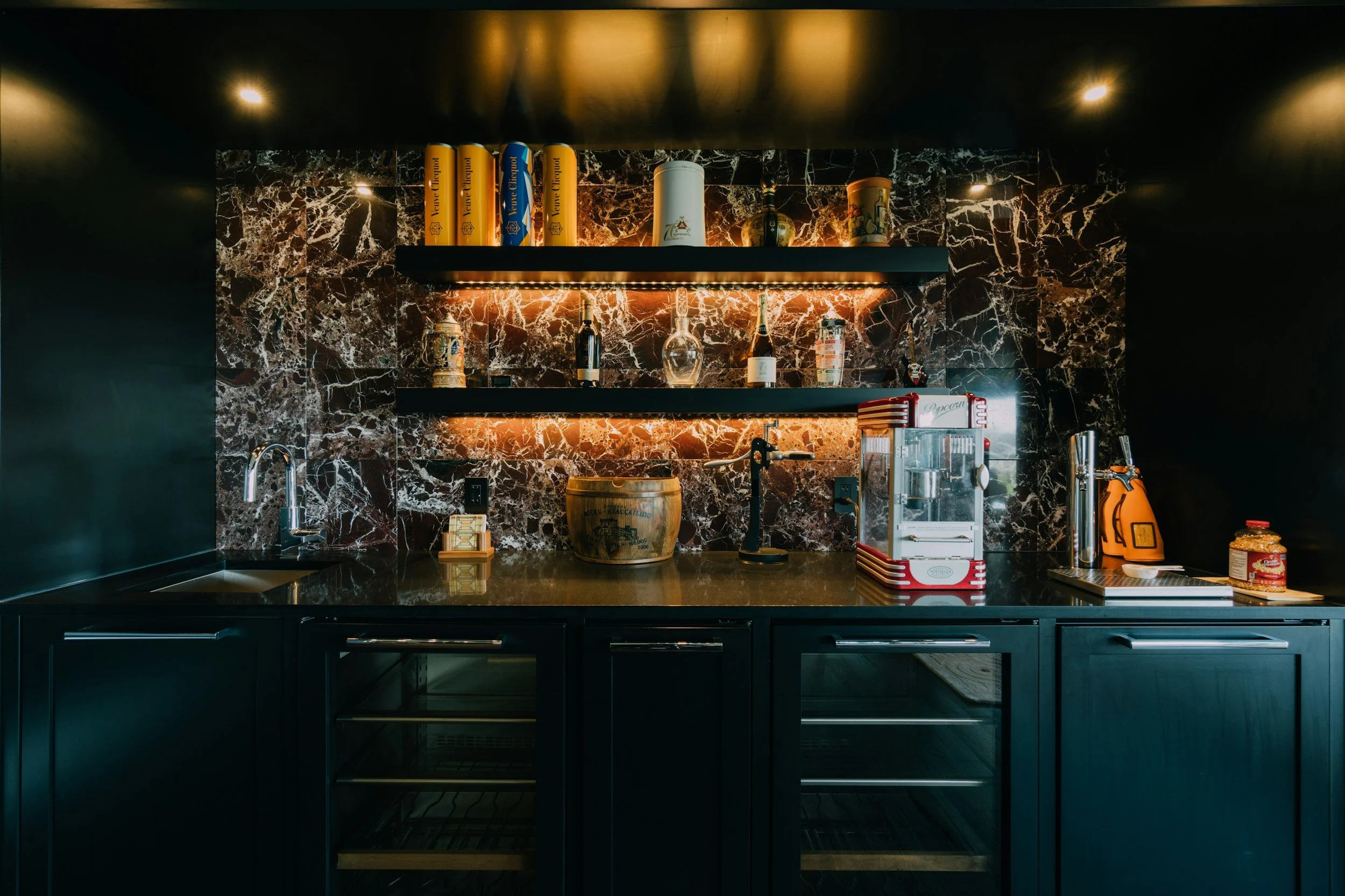 Modern kitchen with black cabinetry, marble backsplash, and illuminated shelves holding books, glassware, and bottles.