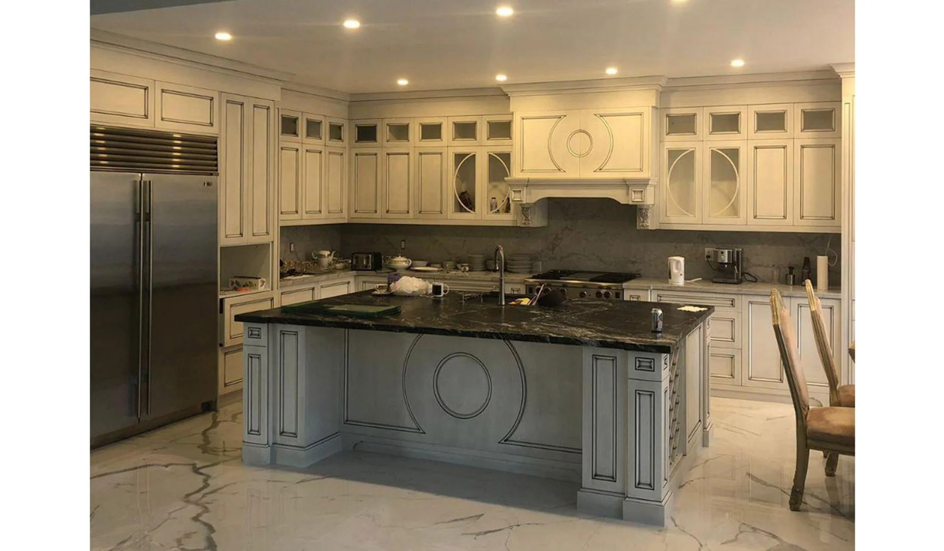 Luxury kitchen with cream-colored cabinets, black marble countertops, and stainless steel appliances, including a double-door refrigerator. An island with similar cream cabinetry and a dark marble top is in the center. There are chairs with beige cushions on the right. The floor is polished marble with brown veins.