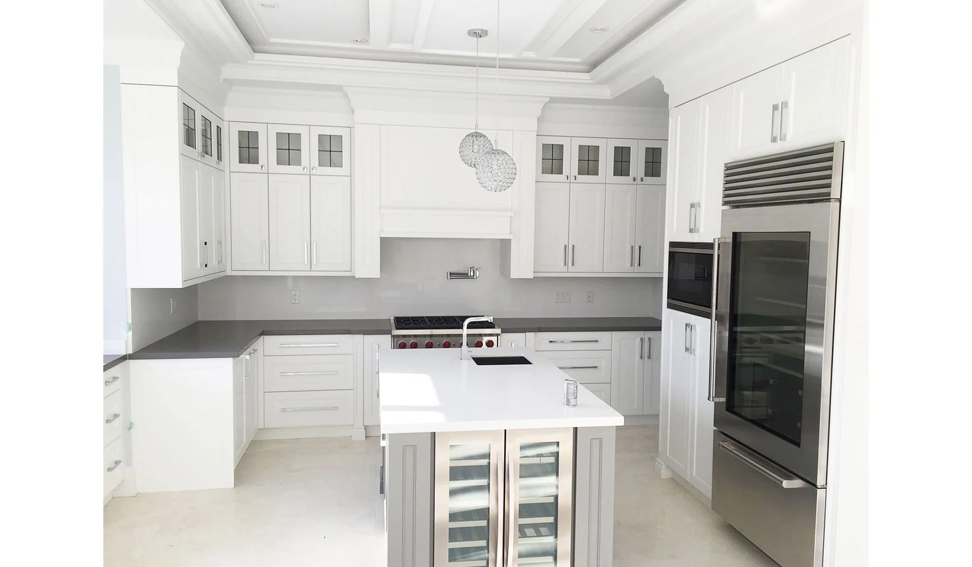 Modern white kitchen with stainless steel appliances, including a refrigerator and microwave, white cabinetry, a white kitchen island with wine storage, gray countertops, a stovetop, and decorative hanging light fixtures.