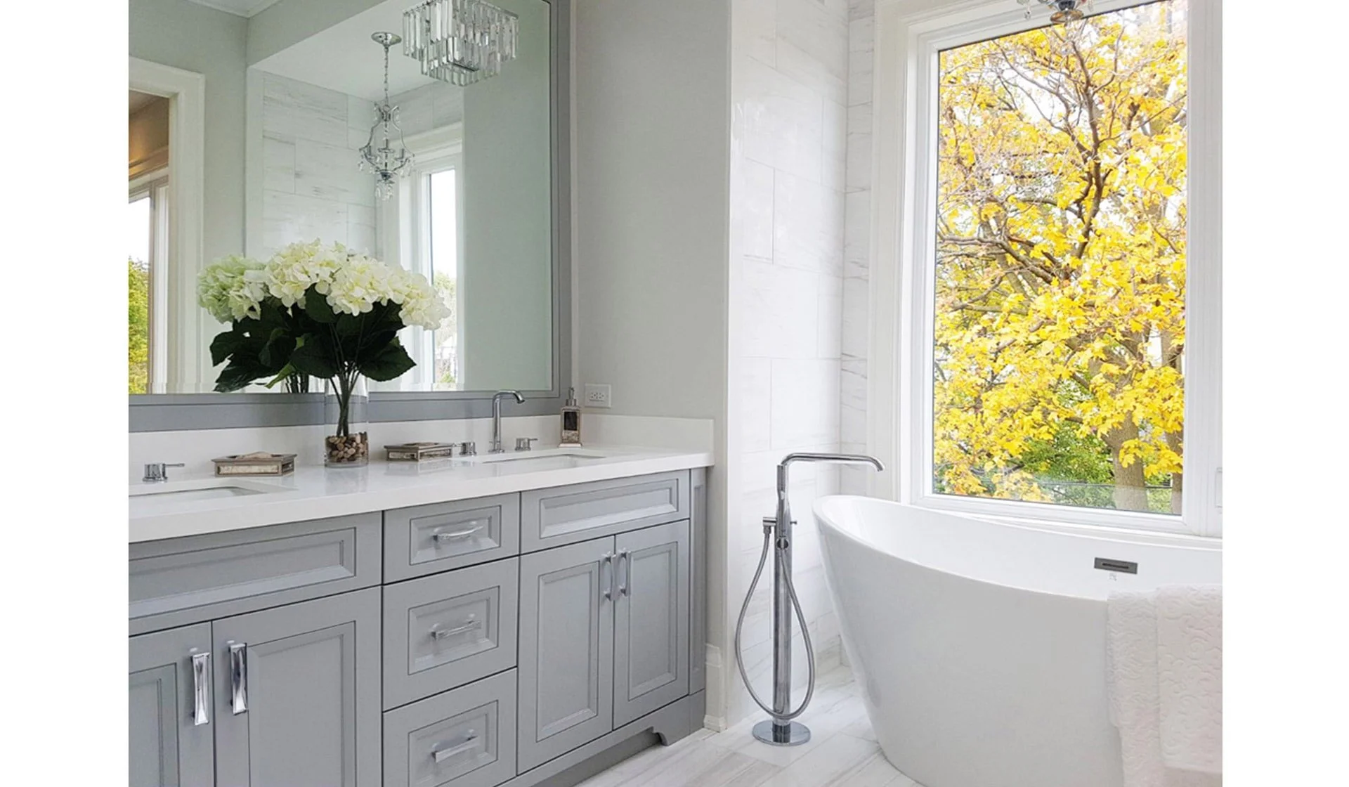 A bright bathroom with a gray vanity, large mirror, white freestanding bathtub, and large window with a view of fall foliage.