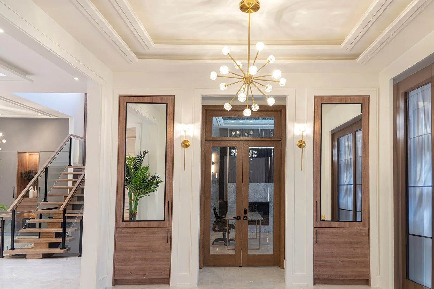 Interior view of a modern home entryway with a gold chandelier, wall sconces, wooden and glass doors, mirrors, and a staircase with a glass and wood railing.