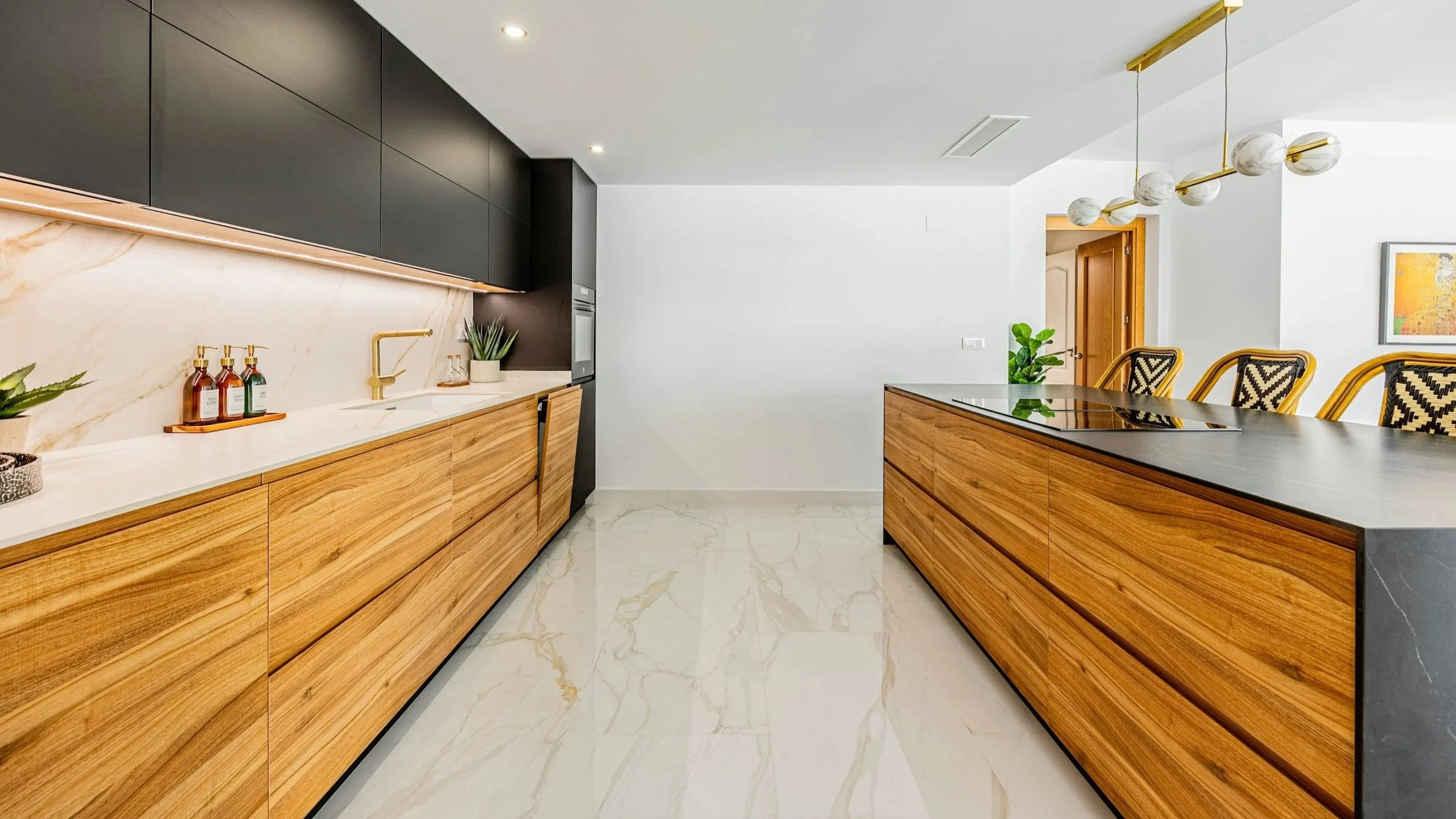 Modern kitchen with black upper cabinets, wooden lower cabinets, white marble backsplash, white marble flooring, and a black countertop island. There are potted plants, soap dispensers, and a gold faucet. A light fixture with marble globes hangs above the island, and a wooden door is visible in the background.