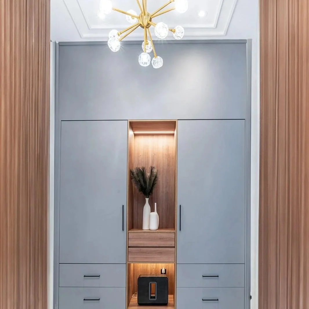 A modern light gray wardrobe featuring a built-in wooden display area with vases and decorative feathers, set against wood-paneled walls and illuminated by a contemporary gold chandelier with multiple glass bulbs.