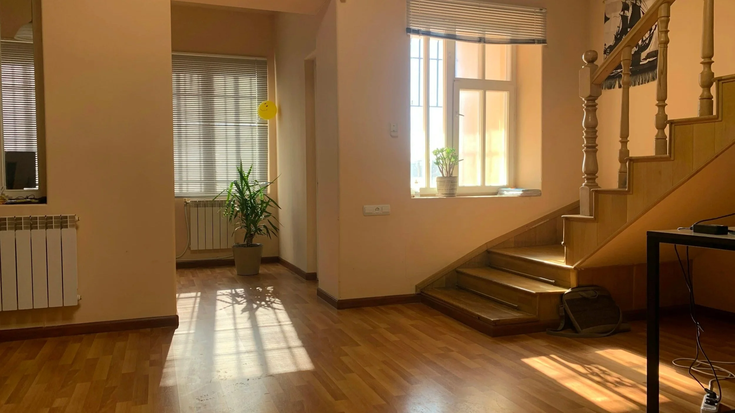 Interior of a house with wooden floors, a staircase with wooden banisters, a large window with blinds, a potted plant near the window, and sunlight streaming in.