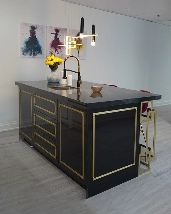 Modern kitchen island with a black and gold finish, a built-in sink, and hanging pendant lights. Artworks are displayed on the wall behind, and a vase with yellow flowers sits on the island.