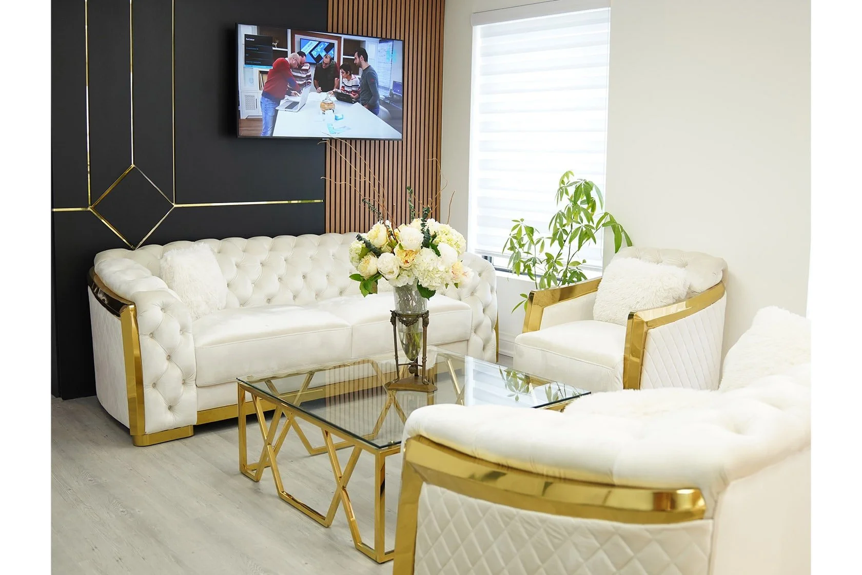 Elegant waiting area with white tufted sofas and armchairs with gold accents, a glass-topped coffee table, a vase of white flowers, a green plant, and a wall-mounted TV showing a meeting scene, all in a modern, well-lit room.