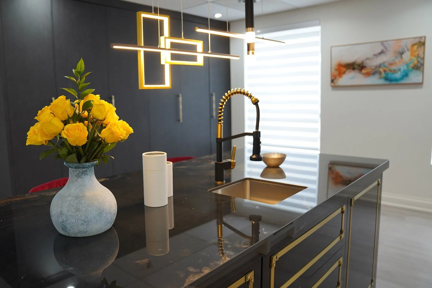 Modern kitchen with black marble island, yellow roses in a gray vase, white canisters, and black matte faucet with a gold flexible hose. Pendant lights hang above, with a window and abstract art on the wall.