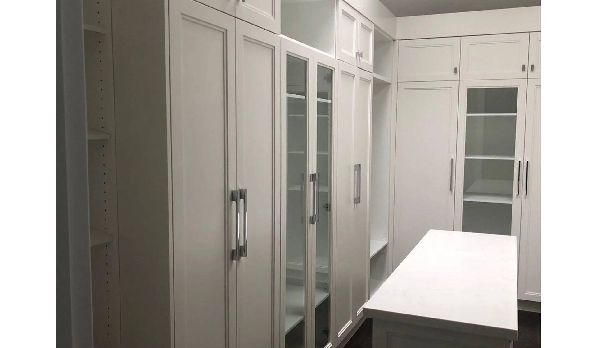 Interior view of a white walk-in closet with multiple cabinets and shelves, including glass-fronted doors and a central island.