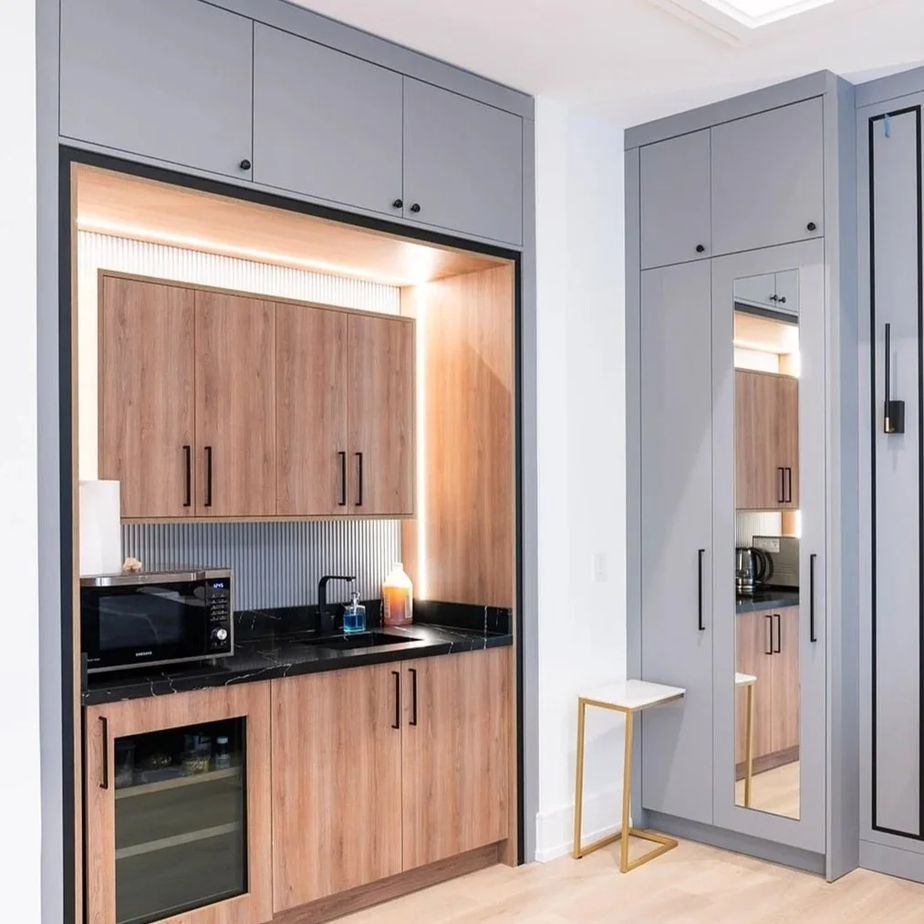 Modern kitchenette with wooden cabinets, black countertop, microwave, small refrigerator, and a sink with a black faucet. Adjacent to a tall grey cabinet with a mirror and a small white and gold side table.