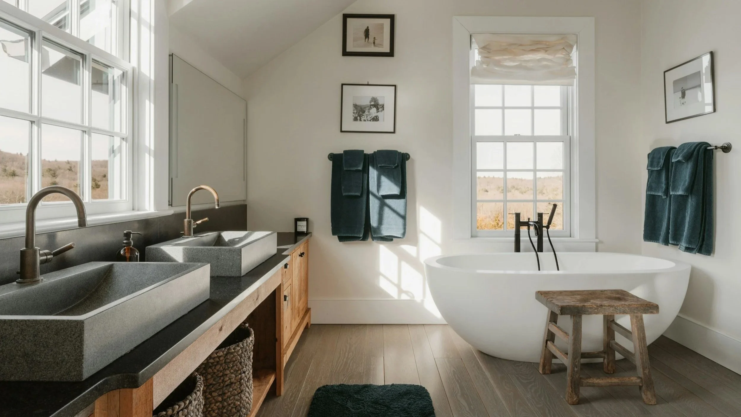 Bright bathroom with double stone sinks, black countertop, wooden cabinet, white bathtub, large window with a scenic outdoor view, teal towels, framed black and white photos, and a small rustic wooden stool.