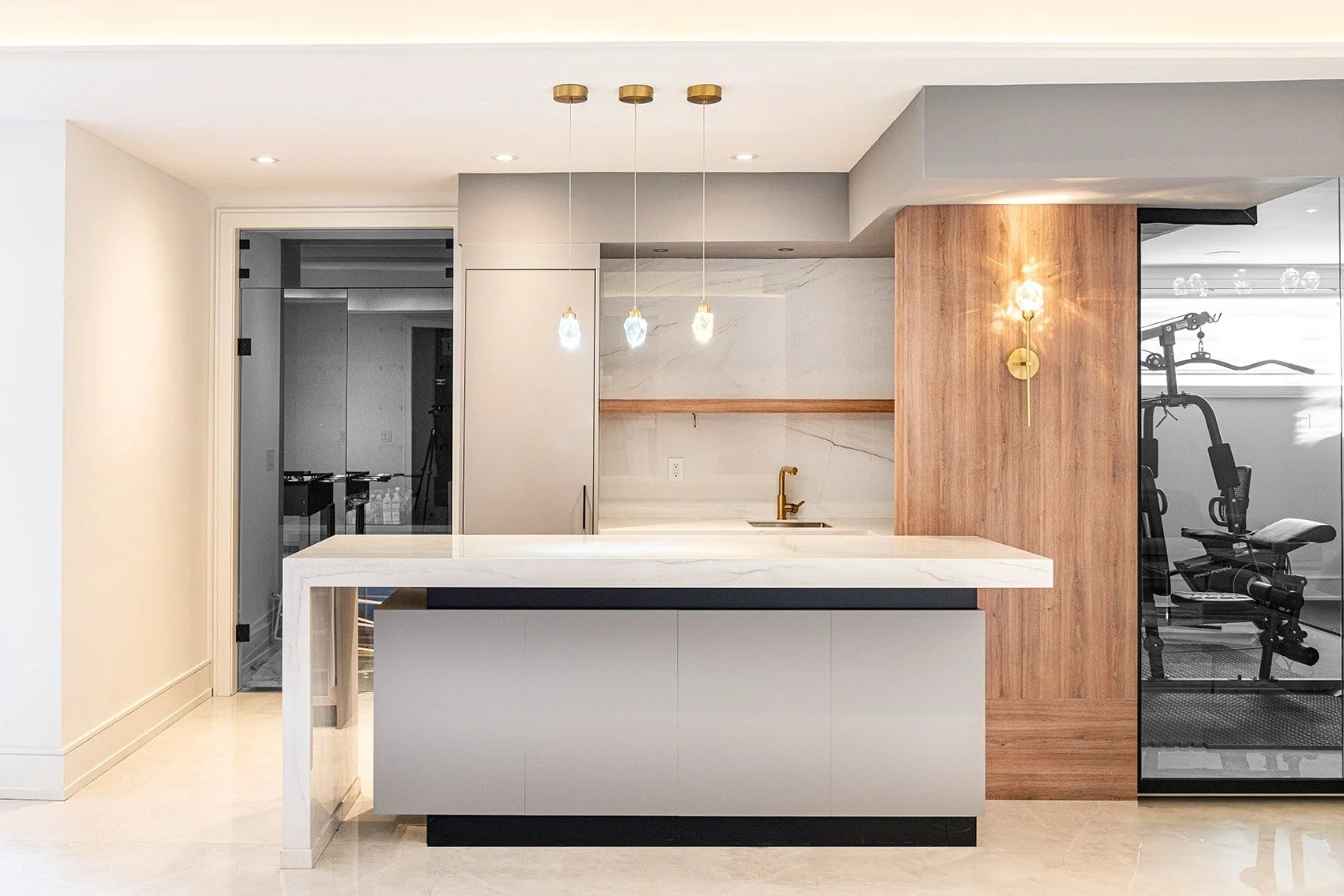 Modern kitchen with white marble island, gold pendant lights, gray cabinetry, and wooden accent wall. Visible fitness equipment behind glass wall.