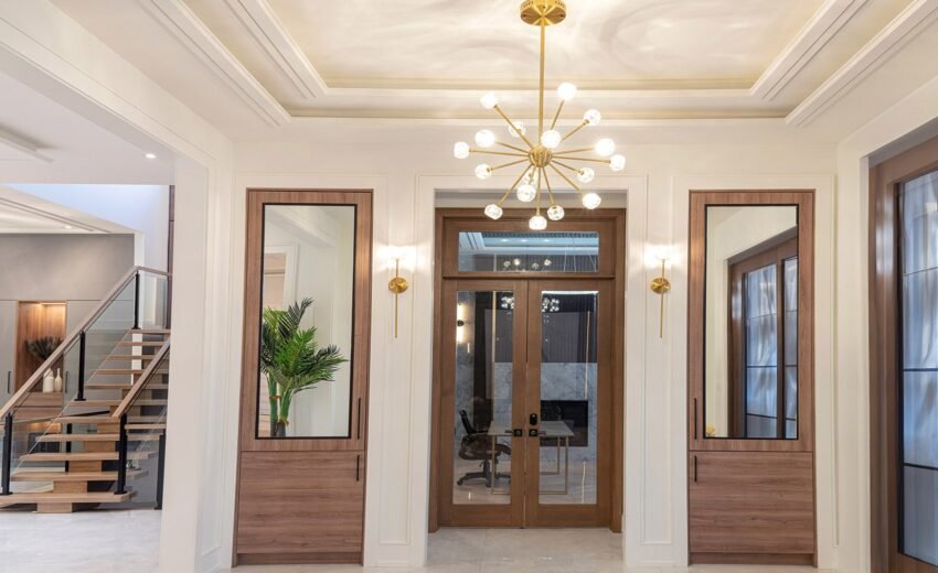 Interior entryway with double glass doors, modern chandelier, wall sconces, wall mirrors, and a staircase visible on the left.