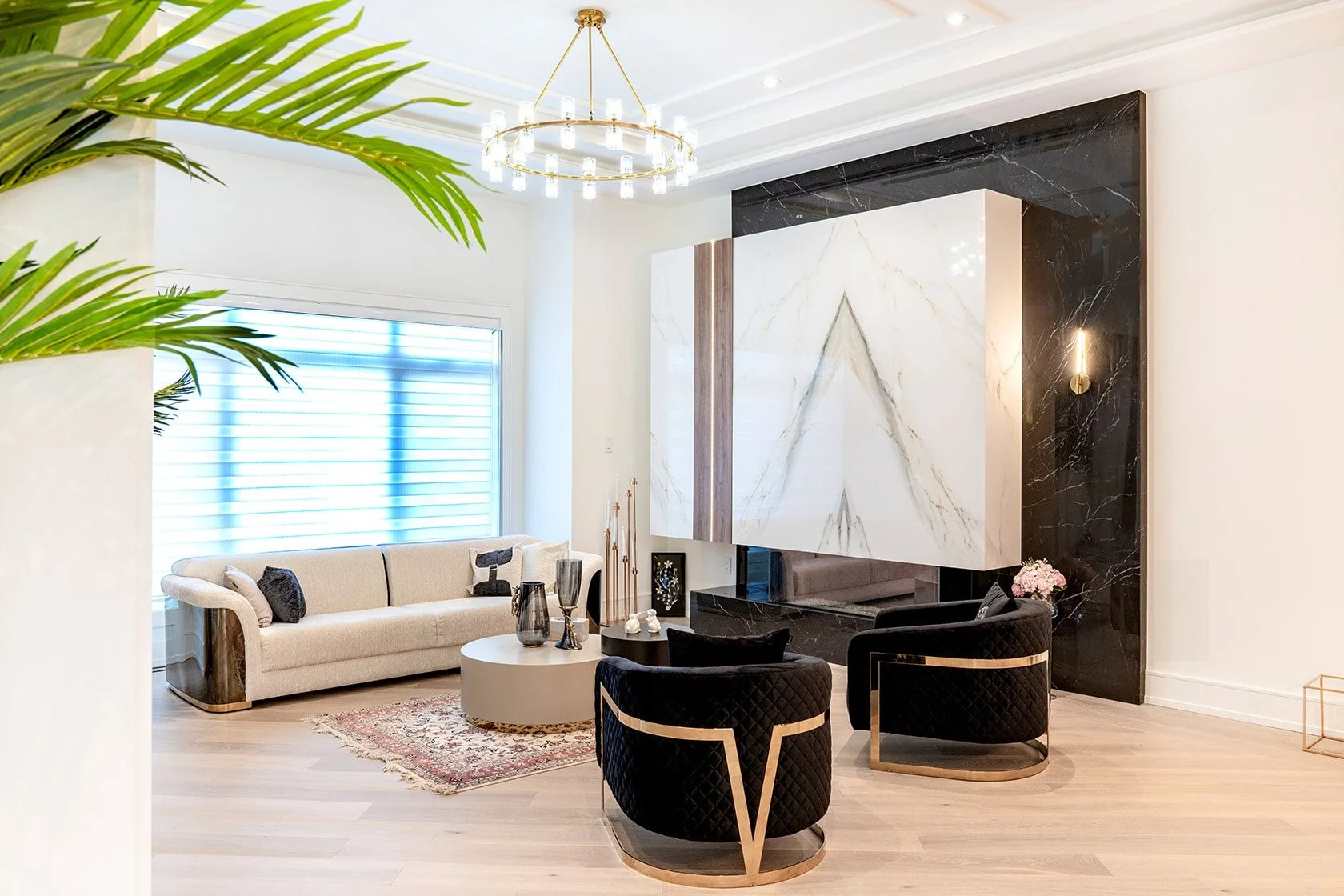 Modern living room with a beige sofa, black velvet armchairs with gold accents, a round coffee table, decorative vases, a patterned rug, a large marble wall feature, and a chandelier.