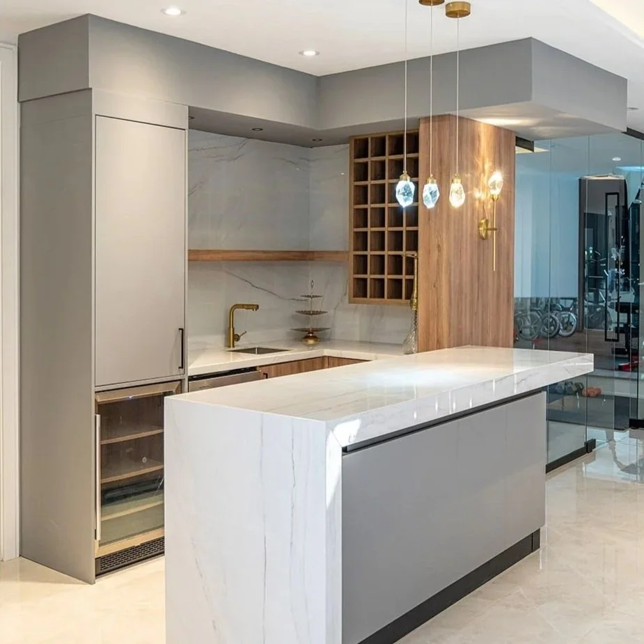 Modern kitchen with white marble island, gray cabinetry, a small wine fridge, gold faucet, and hanging pendant lights, with a wooden accent wall and open shelving.