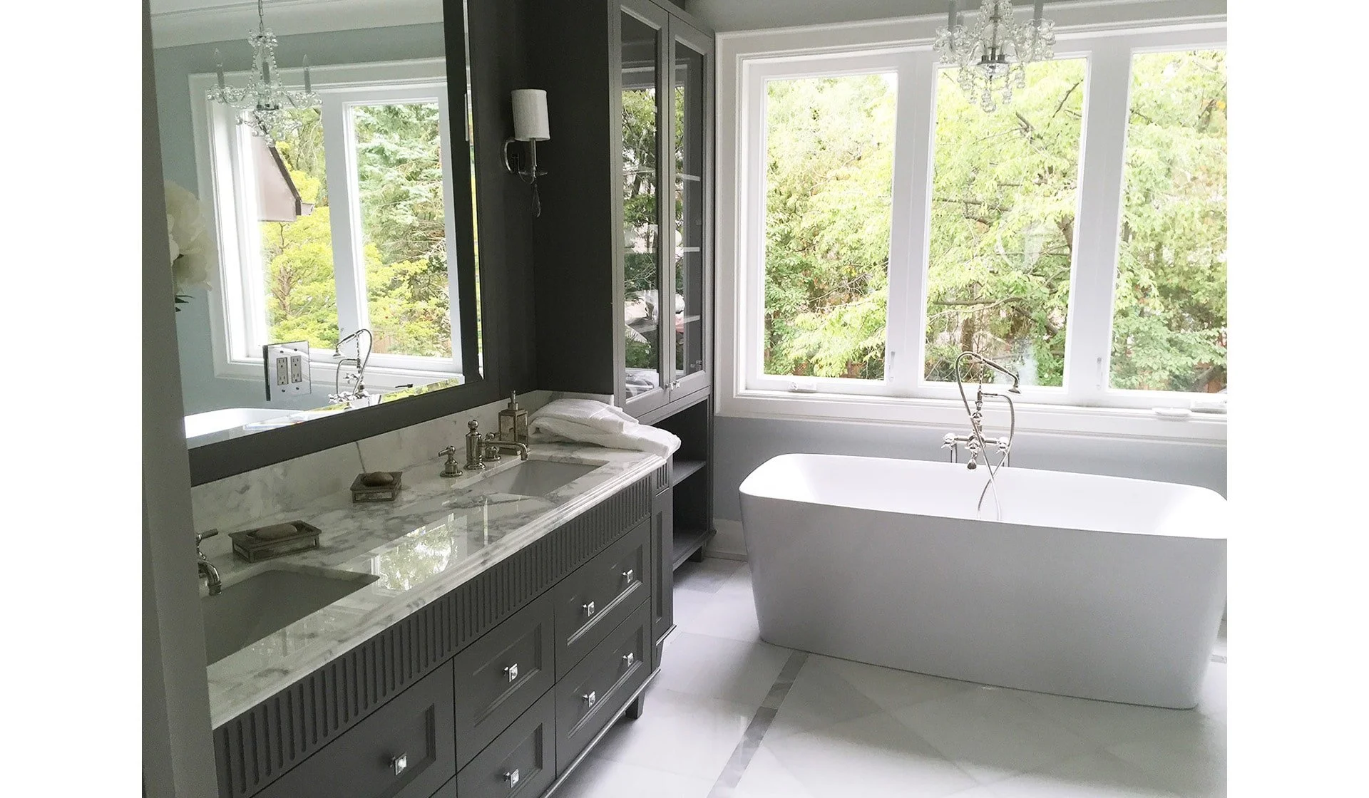 Bathroom with a freestanding white bathtub near large windows with trees outside, a marble-topped gray vanity with multiple drawers and a mirror, and a chandelier hanging from the ceiling.
