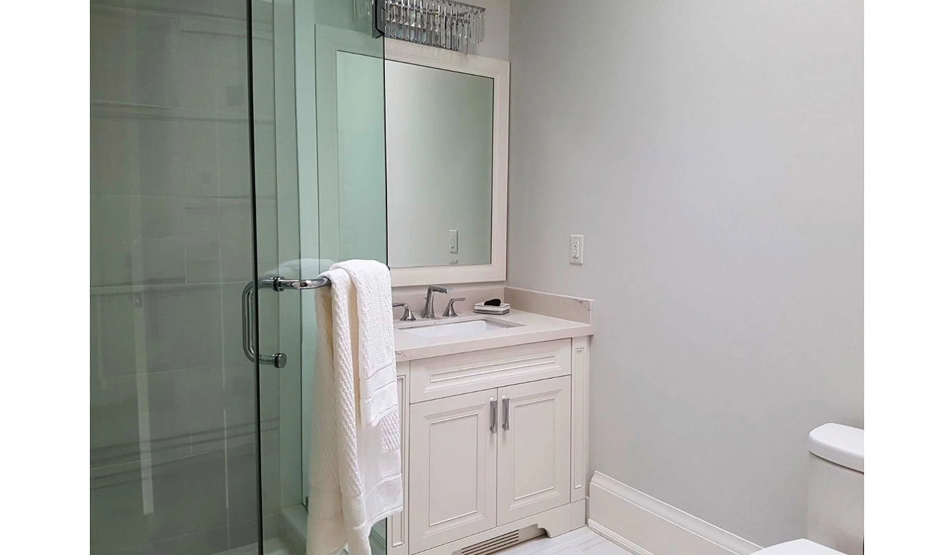 Bathroom with a white vanity, sink, and silver faucet, large mirror, glass shower door, towel hanging on the door handle, and a toilet in the corner, painted in light gray.