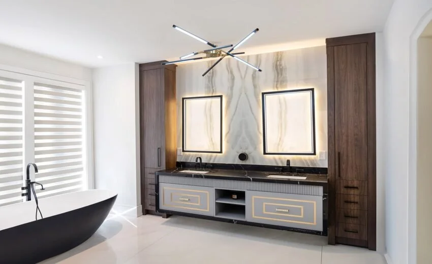 Modern bathroom with a freestanding black bathtub, a large vanity with two sinks, two illuminated mirrors, and dark wood cabinetry. There is a stylish black and gold chandelier overhead and a window with white horizontal blinds.