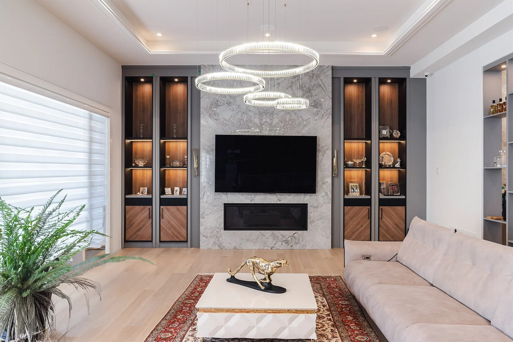 Modern living room with a white wall, built-in wooden and gray shelving units, a large flat-screen TV, and a rectangular fireplace underneath. Three circular chandeliers hang from the ceiling, and there is a beige sofa and a coffee table with a decorative gold sculpture. A patterned area rug and a large window with white blinds are also visible.