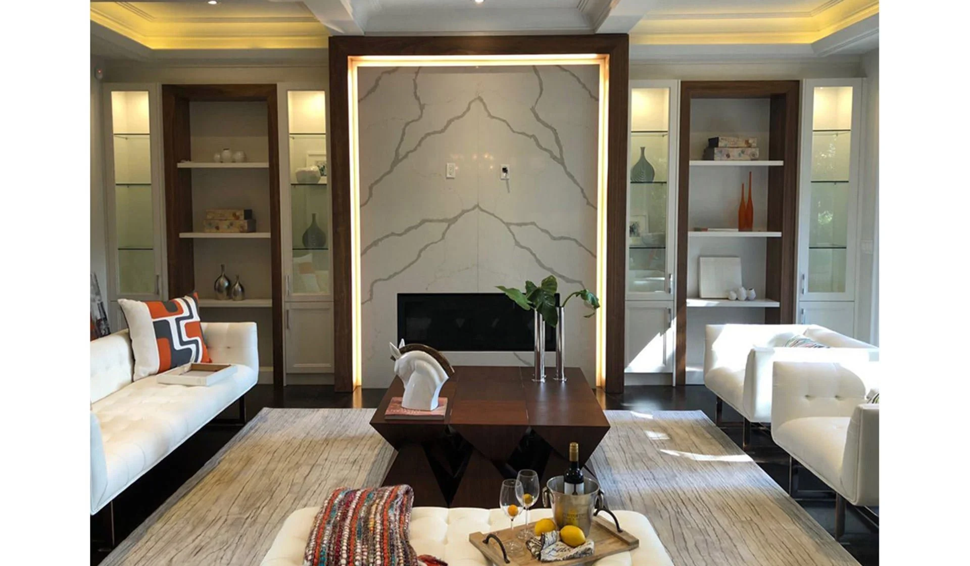 Living room with white sofas, a dark wooden coffee table, built-in shelves with decorative items, a fireplace, and a beige rug.
