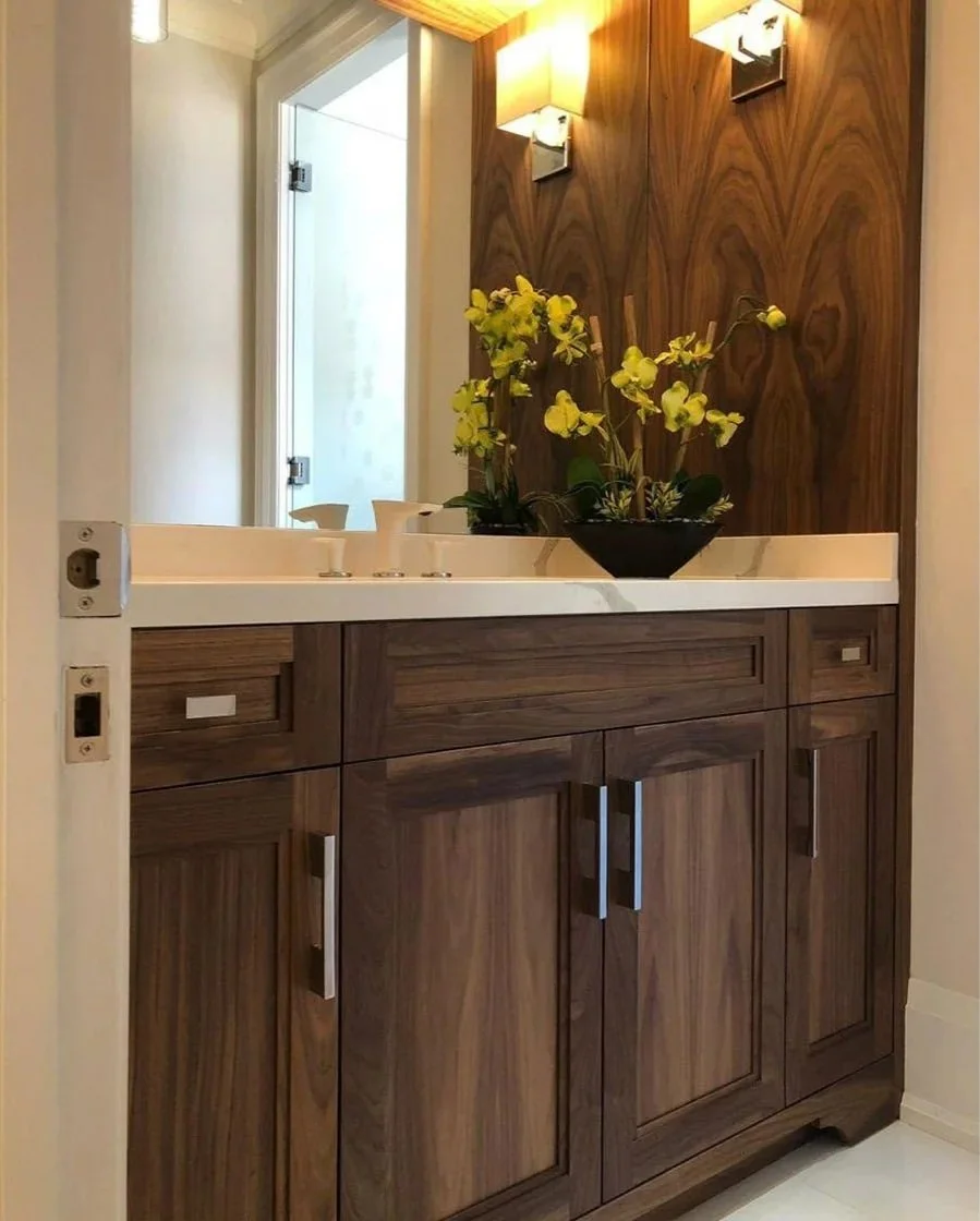 Bathroom vanity with a wooden cabinet, white countertop, and a black vase with yellow flowers. There is a wall-mounted light fixture above and a mirror reflecting a frosted glass door.