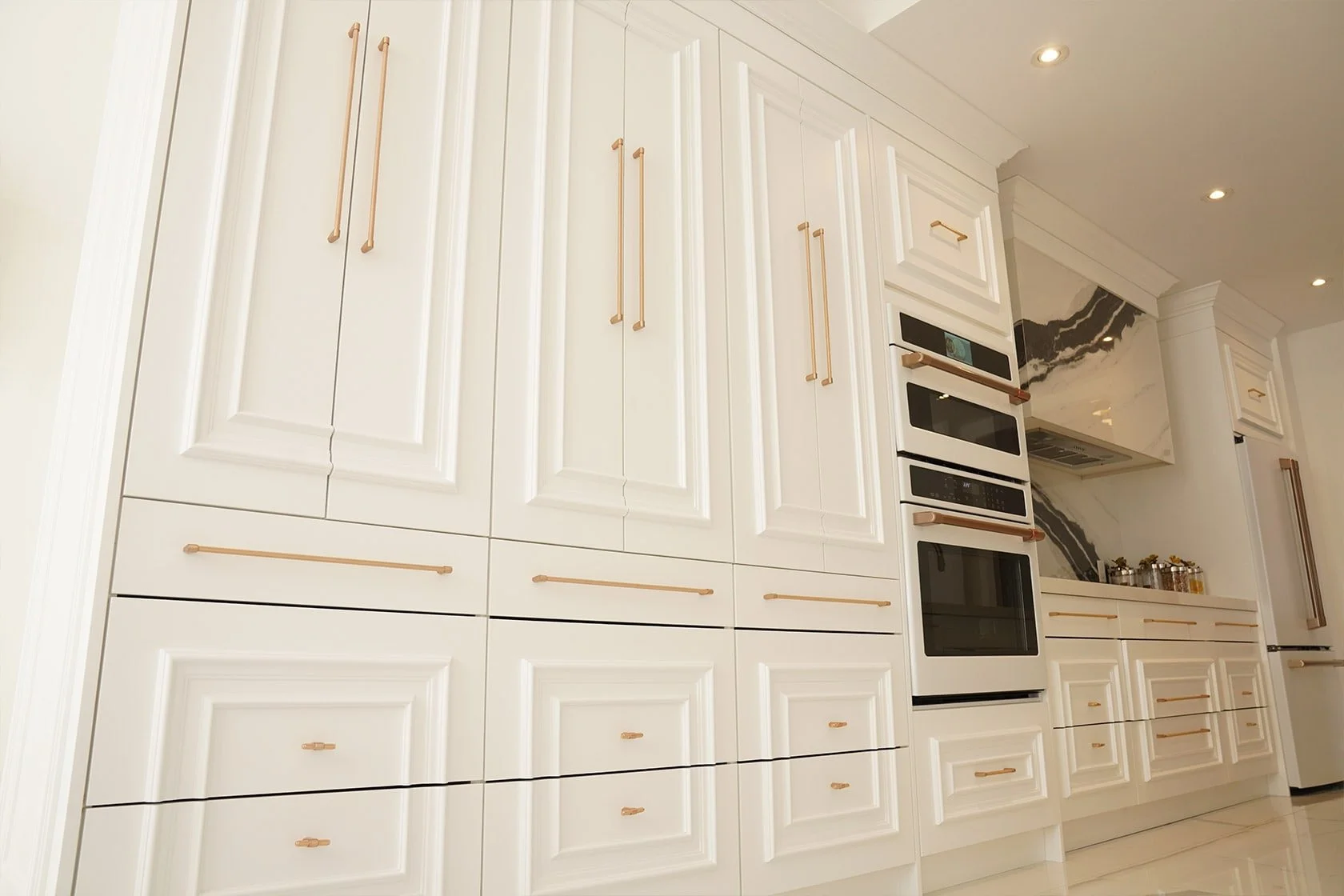 Modern kitchen with white cabinets, gold handles, and black and gold appliances, marble backsplash, and recessed lighting.